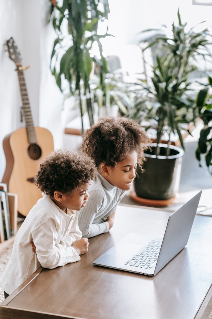 Two children focused on a laptop at home, surrounded by plants and a guitar, exemplifying digital learning.